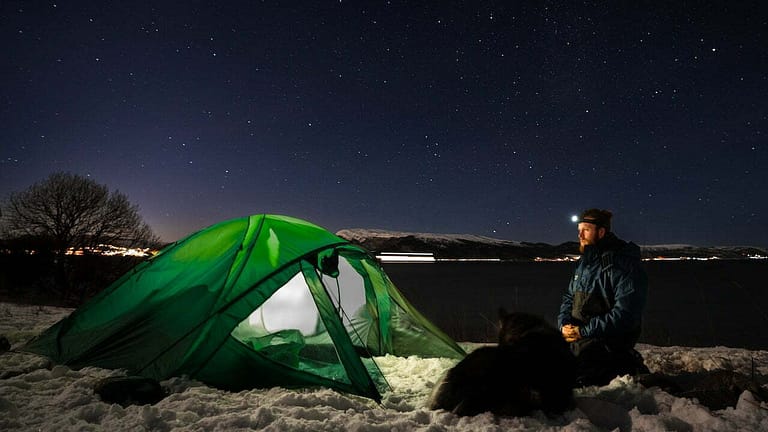 A man sits in the snow near a tent and a dog